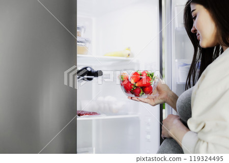 Thoughtful hungry young pregnant woman standing in front of open fridge, takes strawberry from refrigerator in middle of night at home in kitchen, closeup. Healthy eating, nutrition, pregnancy diet Thoughtful hungry young pregnant woman standing in front of open fridge, takes strawberry from refrigerator in middle of night at home in kitchen, closeup. Healthy eating, nutrition, pregnancy diet 119324495