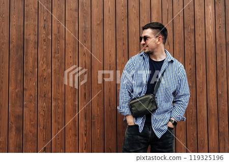 A man in a blue checkered shirt smiles while standing outside a modern building on a sunny day 119325196