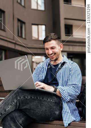 Young man working on a laptop while seated on a bench outside 119325294