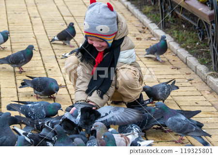 Cute little boy in green rubber boots is feeding pigeons from the bench. Image with toning and selective focus Cute little boy in green rubber boots is feeding pigeons from the bench. Image with toning and selective focus 119325801
