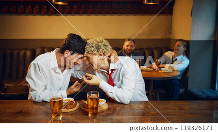 Two colleagues share heartfelt laugh at modern bar, leaning in close, bonding over drinks and snacks in casual setting. Two colleagues share heartfelt laugh at modern bar, leaning in close, bonding over drinks and snacks in casual setting. 119326171