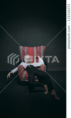Reclining in colorful armchair, man in historical attire listening music in modern headphones, symbolizing relaxation against vintage background. Reclining in colorful armchair, man in historical attire listening music in modern headphones, symbolizing relaxation against vintage background. 119326253