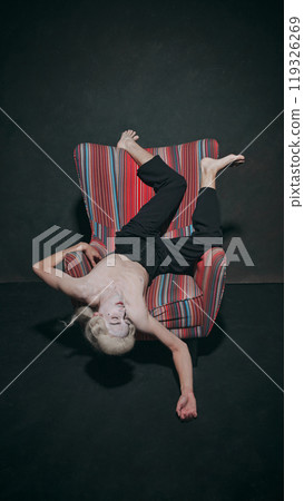 Young man lying on armchair in upside-down pose against vintage style studio background. Blurring lines between past and present Young man lying on armchair in upside-down pose against vintage style studio background. Blurring lines between past and present 119326269
