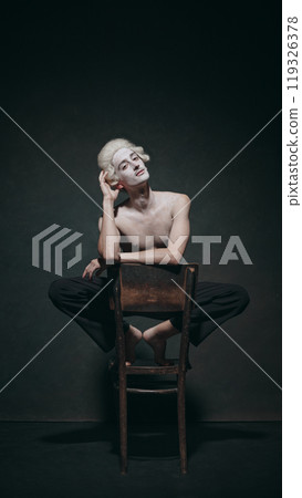 Cross-legged and thoughtful, young man sits elegantly on chair, aristocratic presentation, framed by classic postures in minimalistic style. 119326378