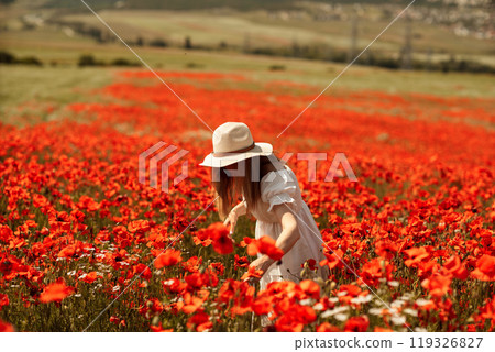 Field poppies woman. Happy woman in a white dress and hat stand with her back a blooming field of poppy. Field of blooming poppies. Field poppies woman. Happy woman in a white dress and hat stand with her back a blooming field of poppy. Field of blooming poppies. 119326827