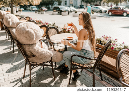 Portrait of happy woman sitting in a cafe outdoor drinking coffee. Woman while relaxing in cafe at table on street, dressed in a white T-shirt and jeans Portrait of happy woman sitting in a cafe outdoor drinking coffee. Woman while relaxing in cafe at table on street, dressed in a white T-shirt and jeans 119326847