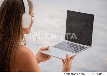A woman is sitting on a beach with a laptop in her lap. She is wearing headphones and she is listening to music. 119326858