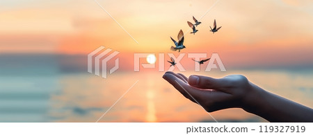 A gentle hand extends towards the horizon, where birds are taking flight against a vibrant sunset over the calm sea. A gentle hand extends towards the horizon, where birds are taking flight against a vibrant sunset over the calm sea. 119327919