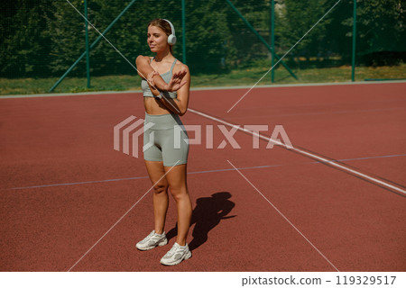 An Active Woman Engaged in Stretching While on a Tennis Court Listening to Music with Headphones 119329517