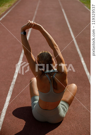 A fit and determined female is stretching her body on a running track, preparing for her workout 119329537