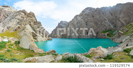 The image shows a blue azure lake surrounded by dark rocks. The water is clear and calm, reflecting the surrounding mountains, creating a magical and fabulous landscape of southern Kyrgyzstan. 119329574