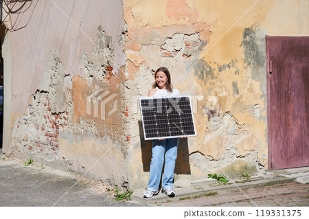 Woman holding solar panel for charging, stands against weathered, textured wall. Smiling female in sunglasses wears white top and blue jeans, standing in sunlit alley in urban scene. Woman holding solar panel for charging, stands against weathered, textured wall. Smiling female in sunglasses wears white top and blue jeans, standing in sunlit alley in urban scene. 119331375