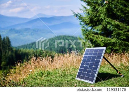 Solar panel on grassy hillside, soaking up sunlight against backdrop of rolling mountains and lush forests. Integration of sustainable energy practices within beauty of nature. 119331376
