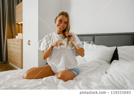 A Joyful Young Woman Taking Care of Her Hair While Sitting on the Bed in a Modern Room 119331693