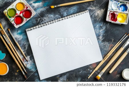 A blank sketchpad surrounded by vibrant watercolor paints, brushes, and palette on a textured background, representing an artist's creative workspace 119332176