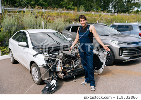 Confused automotive mechanic male in blue coveralls standing by a white car with serious damage after accident, showing scale of damage that needs to be repaired. Concept of car service, repair Confused automotive mechanic male in blue coveralls standing by a white car with serious damage after accident, showing scale of damage that needs to be repaired. Concept of car service, repair 119332866