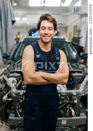 Vertical portrait of positive male car technician wearing uniform smiling looking camera standing posing with crossed hands in auto service shop. Concept of professional automobile maintenance. Vertical portrait of positive male car technician wearing uniform smiling looking camera standing posing with crossed hands in auto service shop. Concept of professional automobile maintenance. 119332958