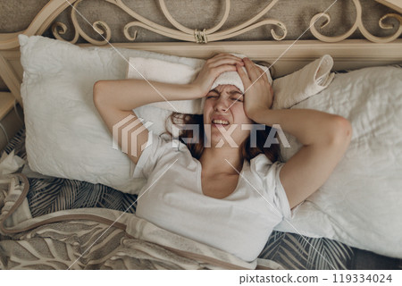 Young woman with headache flu ill sick disease cold at home indoor lying on bed with cold compress on her head at bedroom home Young woman with headache flu ill sick disease cold at home indoor lying on bed with cold compress on her head at bedroom home 119334024