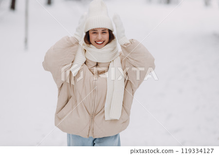 Happy smiling young woman portrait dressed coat scarf hat and mittens enjoys winter weather at snowy winter park Happy smiling young woman portrait dressed coat scarf hat and mittens enjoys winter weather at snowy winter park 119334127