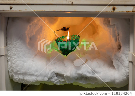 The freezer compartment of the refrigerator is covered with pieces of ice in which there is a burning candle The freezer compartment of the refrigerator is covered with pieces of ice in which there is a burning candle 119334204