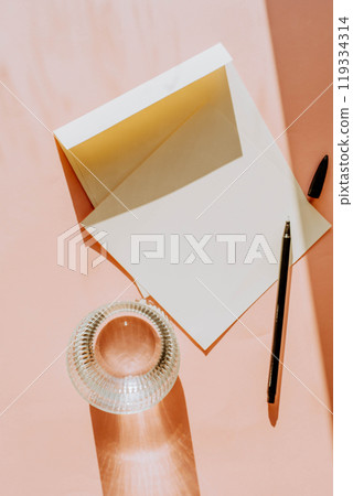 Open envelope and blank paper on a soft, peach-colored surface, accompanied by pen and glass of water 119334314