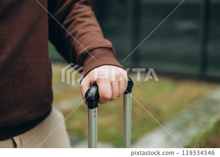 Traveler walking with a suitcase, traveling in the city, cropped view with no face. Passenger waiting at bus station or near hotel. Selective focus on hand holding baggage 119334546
