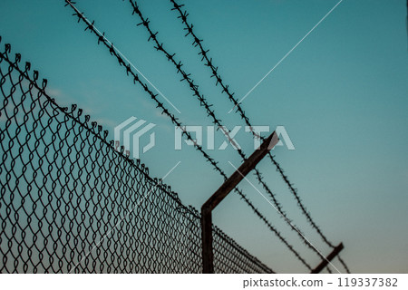 Barbed iron wire against blue sky. Industrial zone protected with a lattice fence. Warehouse, factory territory. Private property. Jail, prison zone.  119337382