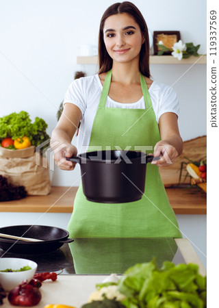 An attractive young dark-haired woman offering ready soup by new keto recipe while standing and smiling in the kitchen. Cooking and householding concepts An attractive young dark-haired woman offering ready soup by new keto recipe while standing and smiling in the kitchen. Cooking and householding concepts 119337569