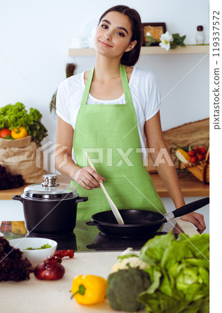 An attractive young dark-haired woman frying meal at worm pan by new keto recipe while standing and smiling in the kitchen. Cooking and householding concepts 119337572