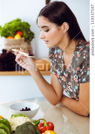 An attractive young dark-haired woman tastes a new recipe for a delicious salad mix while sitting at the table in the kitchen An attractive young dark-haired woman tastes a new recipe for a delicious salad mix while sitting at the table in the kitchen 119337587