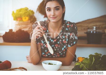 An attractive young dark-haired woman chooses the recipe for a delicious meal while sitting at the table in sunny kitchen An attractive young dark-haired woman chooses the recipe for a delicious meal while sitting at the table in sunny kitchen 119337605