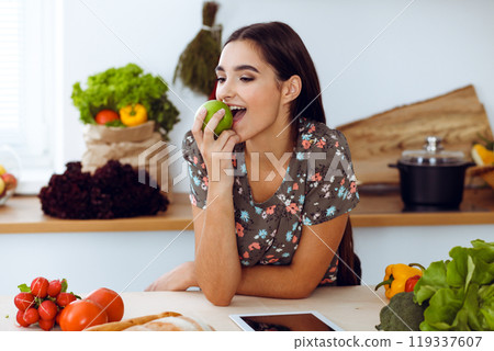 An attractive young dark-haired woman holding green apple and choosing the recipe for a delicious meal while smiling in the kitchen. Tablet pc is the best cookbook 119337607