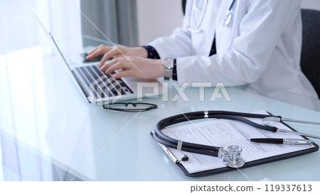Stethoscope and medication history record form are lying on the glass table while doctor is working at the background. Medicine and healthcare Stethoscope and medication history record form are lying on the glass table while doctor is working at the background. Medicine and healthcare 119337613