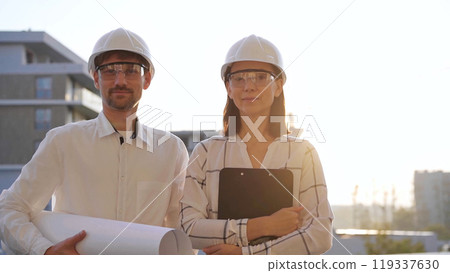 Man and woman architects, wearing safety helmets, are standing near each other and looking at the camera at a construction site during sunrise or sunset. Architecture and engineering concept Man and woman architects, wearing safety helmets, are standing near each other and looking at the camera at a construction site during sunrise or sunset. Architecture and engineering concept 119337630