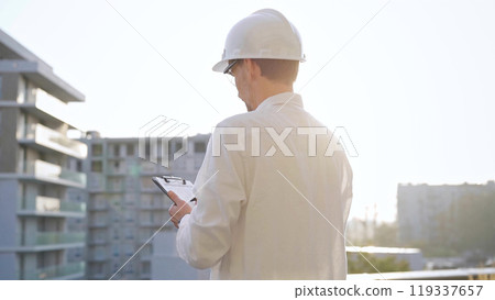 Man construction engineer wearing white shirt and hard hat is making notes on a clipboard while inspecting a building site at sunset, back view. Architecture and engineering Man construction engineer wearing white shirt and hard hat is making notes on a clipboard while inspecting a building site at sunset, back view. Architecture and engineering 119337657