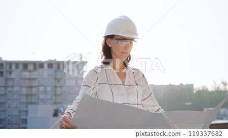 Woman architect, wearing safety helmets and glasses, is holding blueprints and inspecting a building site during sunrise or sunset, front view portrait. Architecture and engineering 119337682
