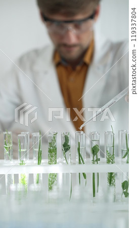 Man scientist wearing a lab coat, white gloves and protective glasses, is dripping liquid from a pipette into a test tube with a green plant inside, vertical portrait. Science and medicine concepts 119337804
