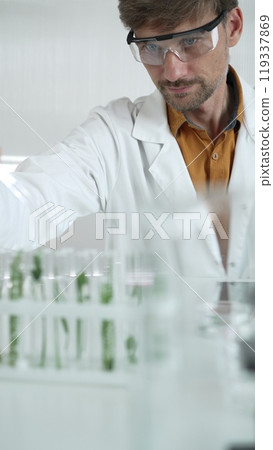 A male science researcher, wearing a lab coat and protective glasses, drips liquid from a pipette into a test tube containing a green plant. His face is focused, captured in a vertical portrait A male science researcher, wearing a lab coat and protective glasses, drips liquid from a pipette into a test tube containing a green plant. His face is focused, captured in a vertical portrait 119337869