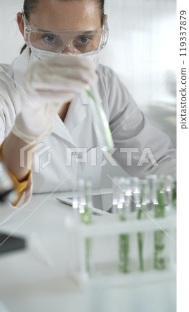 Woman scientist wearing a lab coat, white gloves, face mask and protective glasses, is holding a test tube with plants inside, vertical portrait view. Science and medicine Woman scientist wearing a lab coat, white gloves, face mask and protective glasses, is holding a test tube with plants inside, vertical portrait view. Science and medicine 119337879