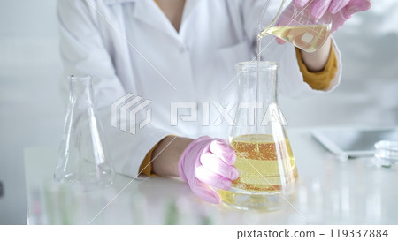 A scientist, wearing a lab coat and pink protective gloves, is pouring a yellow oily liquid from one beaker to another in laboratory, close up. Medicine and science 119337884