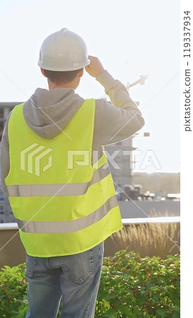 Man engineer with white helmet and safety vest is using tablet computer while inspecting a construction site at sunrise, back vertical view 119337934