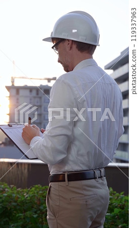 Man a constructive engineer wearing white shirt and hard hat is making notes on a clipboard while inspecting a building site at sunrise, back and vertical view . Architect at work 119337963
