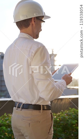 Man a construction engineer wearing white shirt and helmet is using digital tablet while inspecting a building site at sunrise, back vertical view . Architecture concept Man a construction engineer wearing white shirt and helmet is using digital tablet while inspecting a building site at sunrise, back vertical view . Architecture concept 119338054