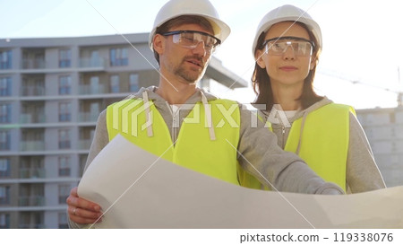 Architects or engineers wearing safety helmets and vests holding blueprint and discussing something on construction site at sunrise, front view. Architecture and engineering Architects or engineers wearing safety helmets and vests holding blueprint and discussing something on construction site at sunrise, front view. Architecture and engineering 119338076
