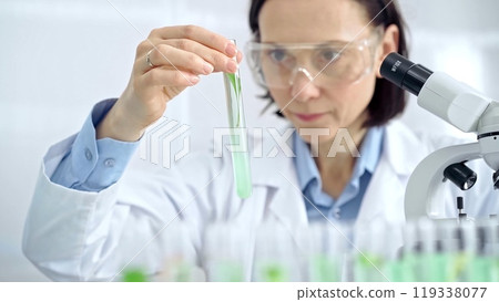 Woman scientist with protective glasses examining lab tube with a green fluid. Microbiology and science concept 119338077