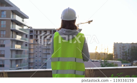 Female construction engineer is taking notes and writing on a clipboard while inspecting a construction site in the early morning at sunrise, back view 119338159