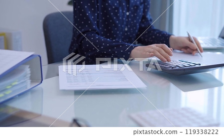 A female accountant with blue dotted blousy is using a laptop computer and calculator to calculate taxes at a glass desk in the office 119338222