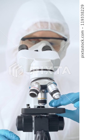 Scientist in ppe analyzing samples with microscope. Close-up of a focused researcher in a lab coat and protective gear using a microscope in a laboratory setting Scientist in ppe analyzing samples with microscope. Close-up of a focused researcher in a lab coat and protective gear using a microscope in a laboratory setting 119338229