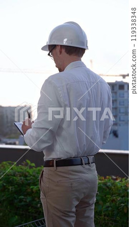 Man a constructive engineer wearing white shirt and hard hat is making notes on a clipboard while inspecting a building site at sunrise, back and vertical view . Architect at work Man a constructive engineer wearing white shirt and hard hat is making notes on a clipboard while inspecting a building site at sunrise, back and vertical view . Architect at work 119338348