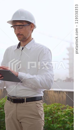 Male construction engineer or architect, wearing a white shirt and helmet, is using a digital tablet while inspecting a building site in foggy weather, front view. Architecture concept 119338353